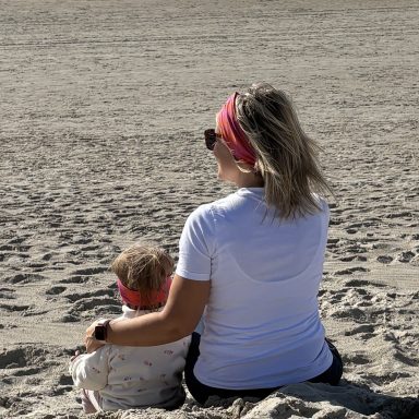 Frau mit Sonnenbrille und Kopftuch sitzt am Strand neben einem Kleinkind.
