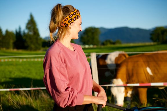 Junge Frau mit Stirnband steht auf einer Wiese, Kühe im Hintergrund.