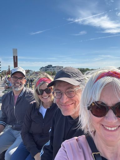 Vier Personen lächeln in die Kamera, vor einer sonnigen Landschaft mit blauem Himmel.