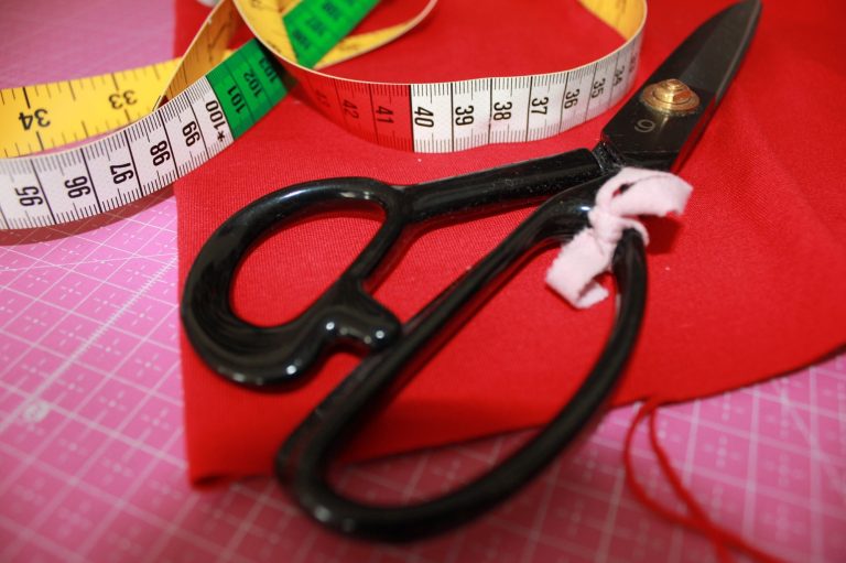 Schneiderschere auf rotem Stoff, umgeben von einem Maßband.